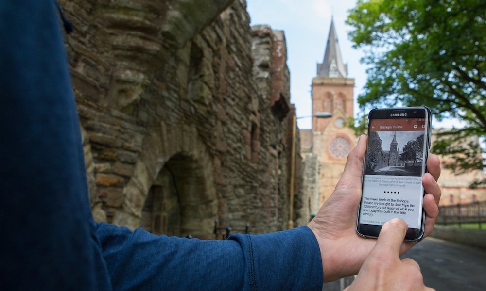 an arm holding a phone near a church with the screens displaying a page form the kirkwall heritage trails app
